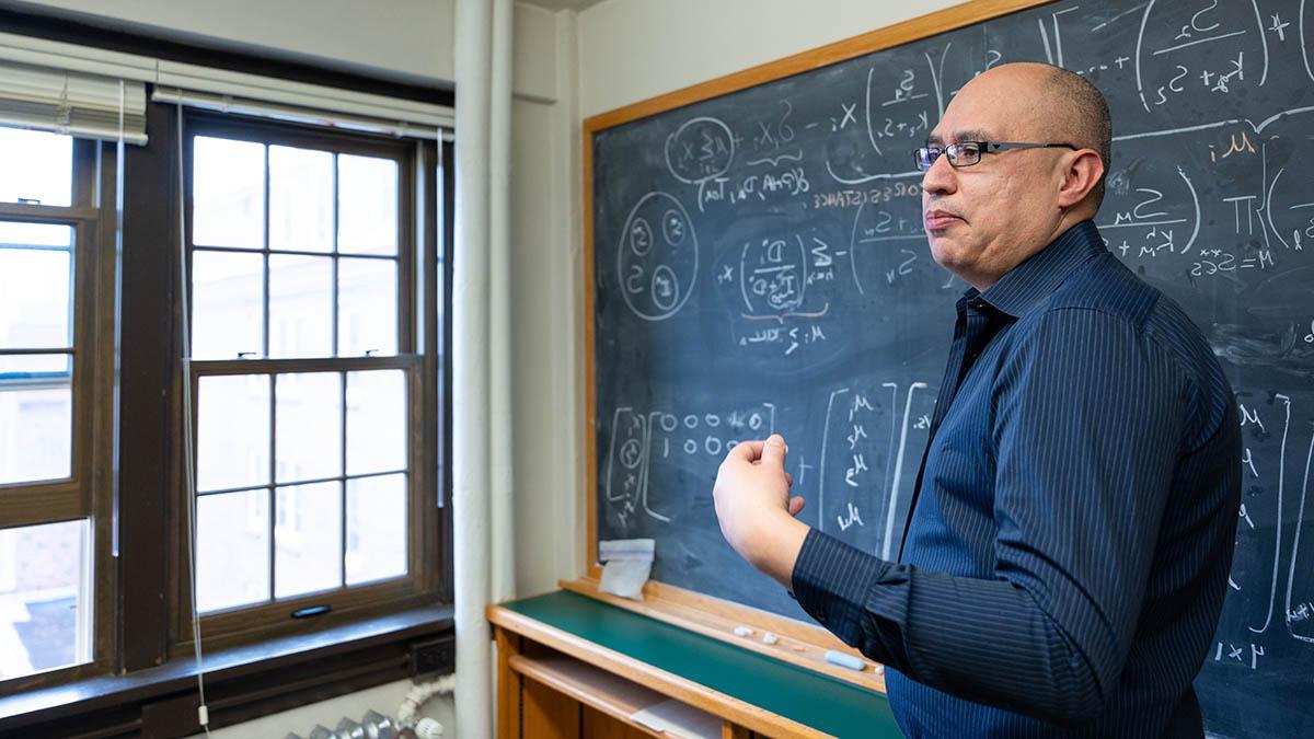 一名戴着眼镜，穿着蓝色衬衫的男子站在黑板上的数学公式前，用手做着动作.